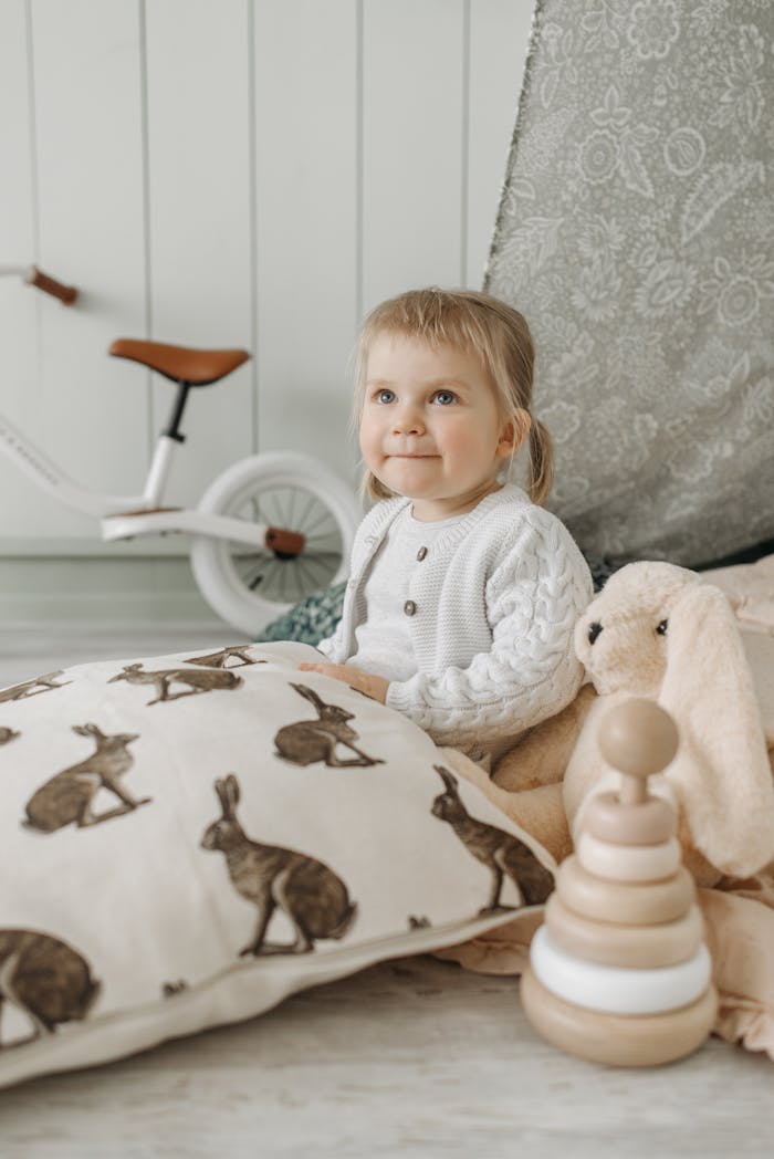 Smiling toddler sitting indoors with toys and a bicycle in view.